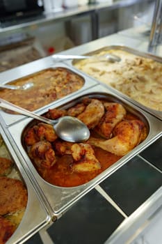 A bustling cafeteria serving line is filled with an array of dishes during a busy lunchtime. Trays of hot food are neatly arranged under the protective glass shield, with customers in the background selecting their meal choices. The variety suggests a diverse menu, catering to different tastes.