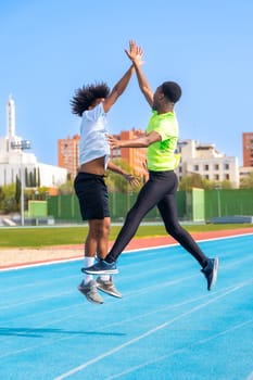 Vertical photo of two african american young friends jumping celebrating and clasping hands in a running track
