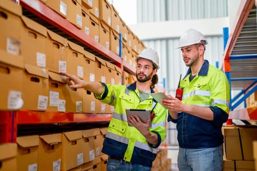Two professional warehouse workers stand between shelves of products with one point to the box and discuss together about work in workplace area.