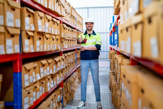 Wide shot of professional warehouse worker man stand with thumbs up and smiling to camera also stay between shelves of product boxes in workplace area.