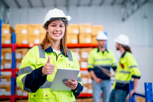 Warehouse worker young woman hold tablet with show thumbs up and look at camera and smiling with her coworkers discuss in the background.