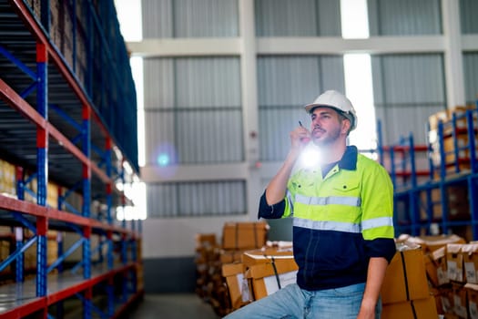Portrait of warehouse worker man use walkie-talkie to contact his co-worker and sit on box and the light shine through his body in workplace area.