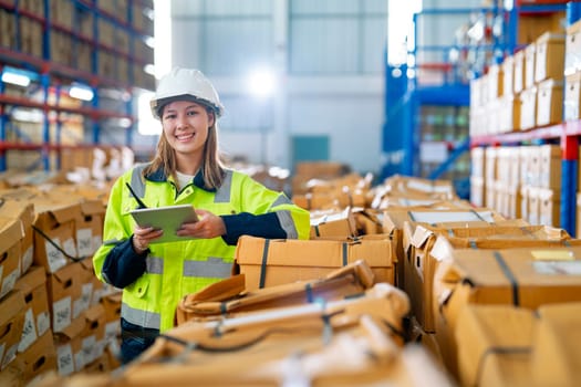 Pretty young warehouse woman hold tablet and stand near stack of product boxes in warehouse workplace and light on the background.