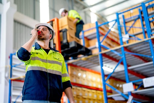 Smart professional warehouse worker use walkie-talkie to contact his co-worker during other worker still carry box or work in the background.