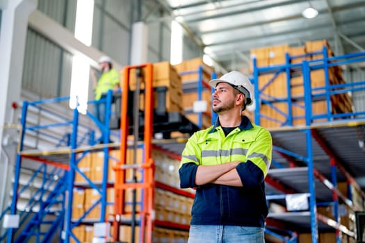 Portrait of Caucasian warehouse worker stand with arm-crossed and look to left side in front of co-worker work and manage boxes on upper layer floor in workplace.