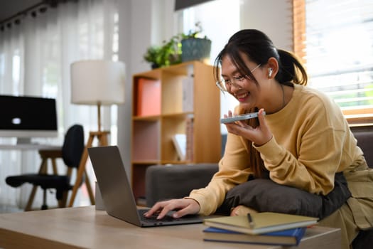 Smiling asian female freelancer having phone conversation and using laptop on sofa.