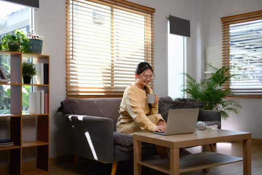 Young woman in casual clothes using laptop sitting on sofa at cozy home.