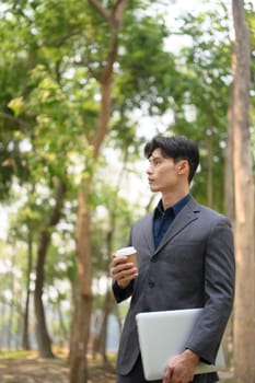 Portrait of handsome businessman holding paper cup of coffee standing in the public park.