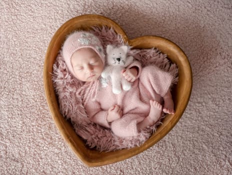 Newborn Girl In Pink Suit With Toy Cat Sleeps In Wooden Heart-Shaped Bowl During Professional Newborn Photoshoot