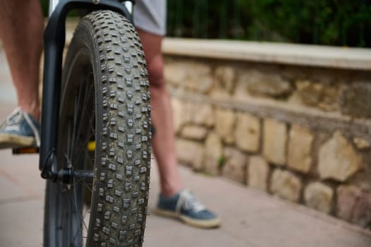 Selective focus on e-bike wheel and feet on pedals of male cyclist in denim sneakers. Shallow tread of a tubeless bicycle tire.