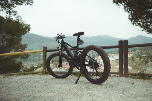 A full size shot of a modern black electric motor bike on the countryside road, parked by a wooden fence, against mountains background in the nature. Copy advertising space. Bike sharing city service.