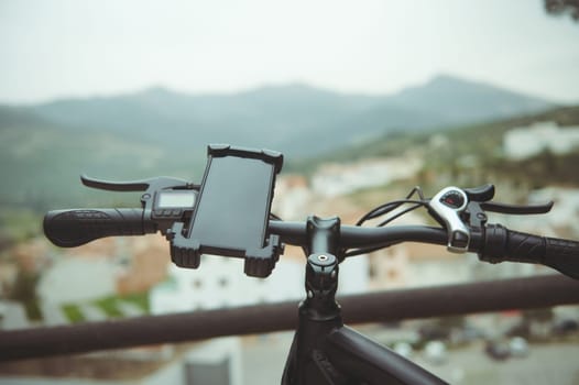 Selective focus on a smartphone fixed on the handlebar of a modern electric bike, with mockup empty black digital touch screen, against the background of blurred medieval city in mountains. Copy space