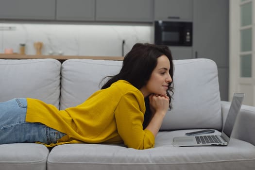 Thoughtful young woman in eyewear using computer while sitting on the sofa at home. High quality photo