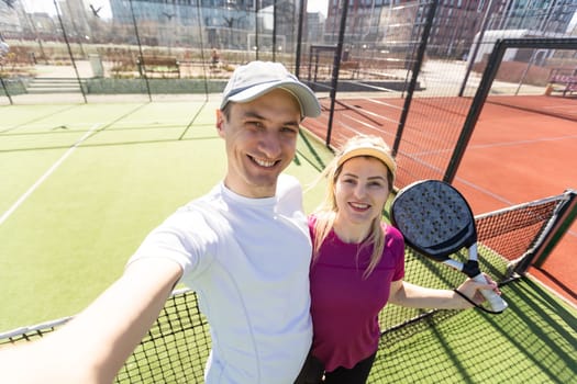 Young couple of tennis players wearing a sportswear holding a racket on their shoulders and a ball in a hand standing outdoor on tennis court at early morning. High quality photo