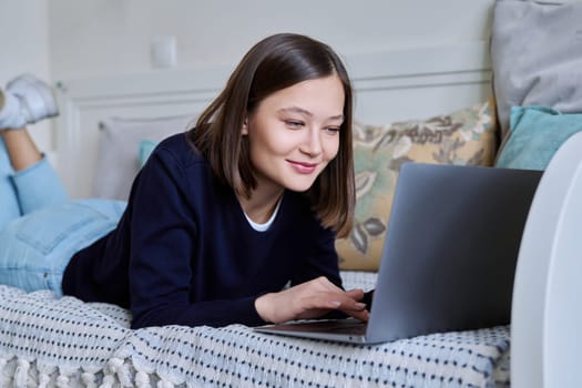 Young woman using laptop computer, typing on keyboard, lying on sofa at home. Internet online technologies for work communication study leisure