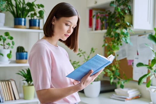 Serious concentrated young woman reading book at home near window. Important study literature guide, interesting fiction, hobby leisure reading