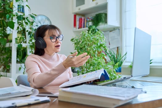 Middle aged woman teacher in headphones with textbook working online using laptop computer in home office. Video conference remote meeting consultation e lesson training education technology concept