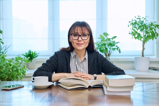 Middle-aged woman reading books, sitting at home at table with pile of books, cup of coffee, relaxed smiling looking at camera. Leisure, lifestyle, literary hobby, mature people concept