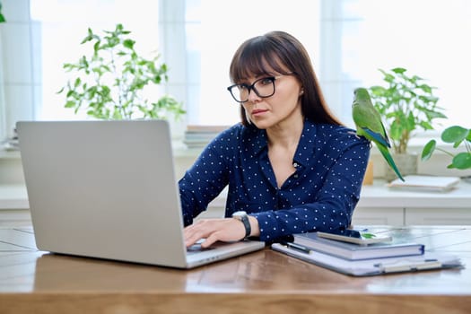 Middle aged woman sitting at workplace with computer in home office, holding pet with green quaker parrot on her shoulder. Work, lifestyle, pets, tropical birds