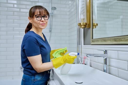 Smiling woman with detergent spray, washcloth cleaning in bathroom, washing sink washbasin. Routine house cleaning, home hygiene, housecleaning service, housekeeping, housework concept