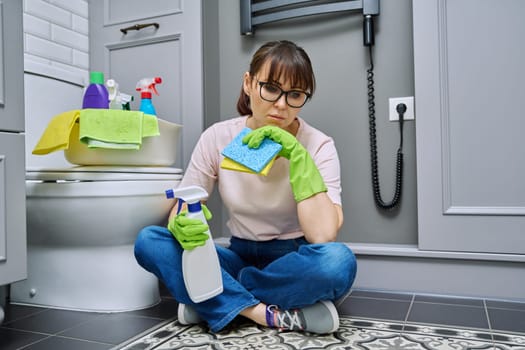 Tired woman sitting on the floor after cleaning bathroom, toilet. Routine house cleaning, home hygiene, housecleaning service, housekeeping, housework