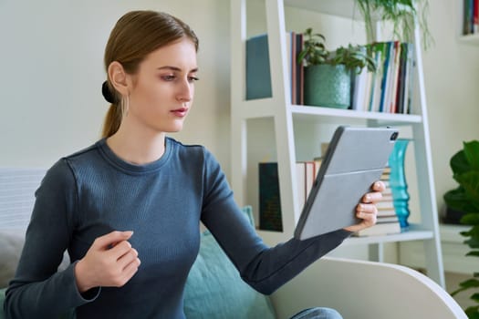 Young teenage female student sitting at home looking talking in web camera of digital tablet. Girl teenager 16,17,18 years old studying remotely, video conference call, e-learning technology education