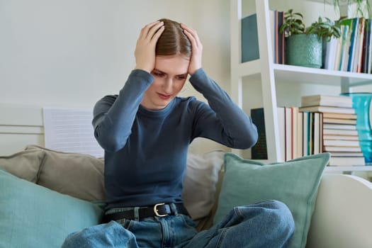 Upset sad lonely young woman sitting on couch at home. Disappointed female 19-20 years old, girl college student in sadness. Depression, stress, mental difficulties, youth concept