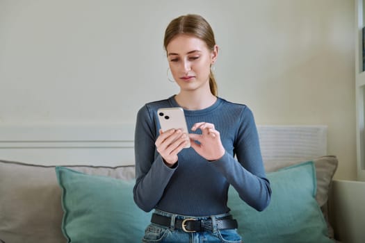 Serious calm female teenager using smartphone, sitting on couch at home, girl 16,17, 18 years old texting reading messages. Modern digital technologies for communication, leisure, learning, shopping