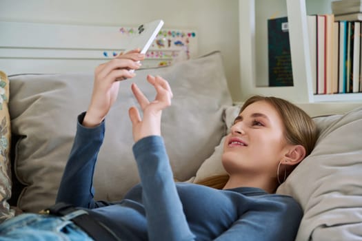 Happy smiling relaxed young female teenager lying on couch at home using smartphone for leisure communication. Youth, lifestyle, digital online technology, gadgets, internet