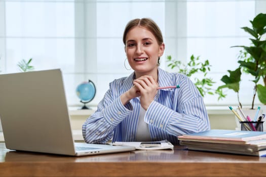 Portrait of high school, college student smiling young female sitting at desk with laptop computer looking at camera. Education, training, e-learning, 16,17,18 year old youth