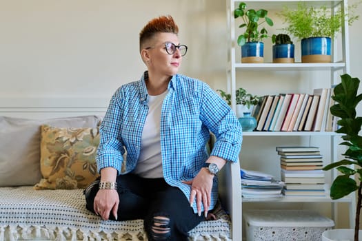 Portrait of middle-aged woman with red hair wearing glasses in casual clothes sitting on couch at home looking out window