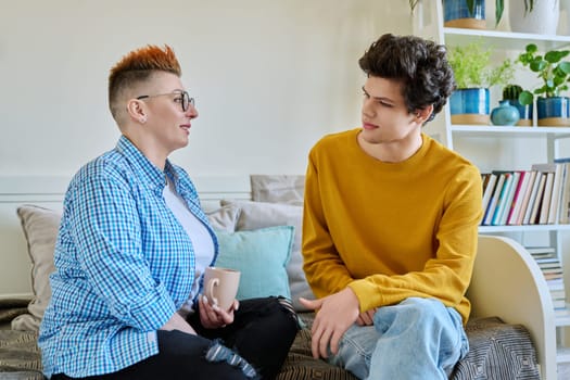 Happy middle-aged mother and son 19-20 years old, talking sitting together on couch at home. Mother's day, lifestyle family communication parenthood tenderness, positive relationship, two generation