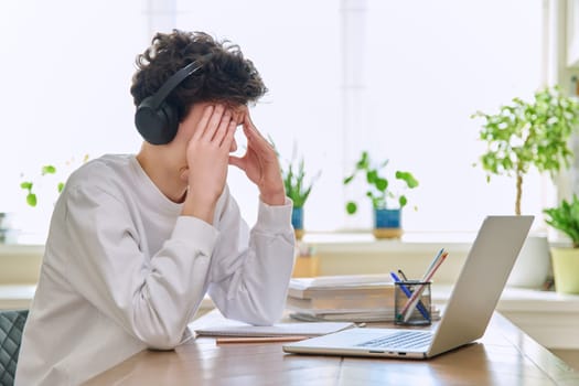 Sad upset young guy student in headphones sitting at home at desk with computer laptop, touching head with hands. Problems headaches troubles in college difficulties in study mental stress depression