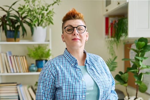 Portrait of smiling middle-aged woman in glasses with red haircut looking at camera in home interior. Mature people, lifestyle, health, life concept