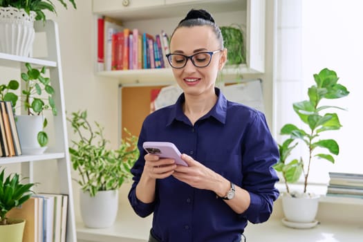 Beautiful smiling woman in her 30s using smartphone, standing in her room at home. Communication, work, leisure, lifestyle, people concept