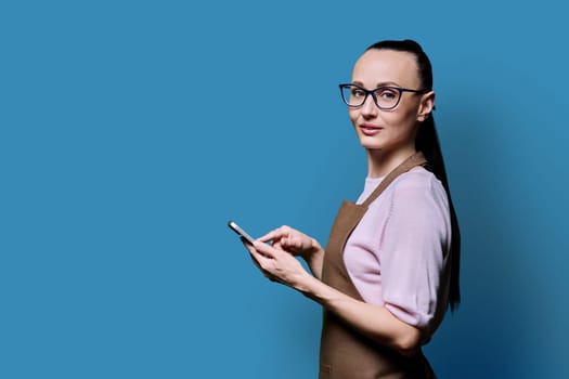 Portrait of 30s woman in apron with smartphone looking at camera on blue background. Female using mobile phone texting receiving sending order, copy space. Technologies applications service small business
