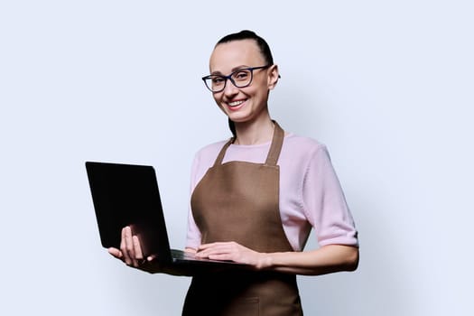 Portrait of 30s smiling happy woman in an apron using laptop, looking at camera on blue studio background. Worker, startup, small business, job, service sector, staff, youth concept
