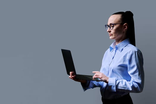 Middle-aged business serious woman using laptop on gray background. 30s successful female teacher mentor manager worker employee director. Internet online technologies, work, electronic teaching