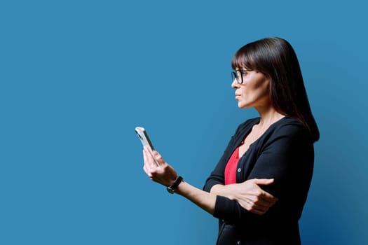 Profile view middle aged serious woman using smartphone on blue background. Mature female looking at phone in hands. Technologies mobile apps applications internet work business leisure communication