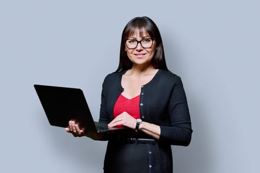 Smiling business middle aged woman using laptop on gray studio background. Confident successful mature female looking at camera. Business, work, job, lifestyle, technology, 40s people concept