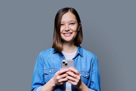 Young happy woman using smartphone in gray background. Smiling 20s female looking at camera texting. Mobile Internet apps applications technologies for work education communication shopping healthcare