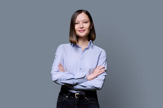 Young confident smiling business woman with crossed arms on grey studio background. Positive happy female in shirt, student worker owner entrepreneur looking at camera. Business work education people