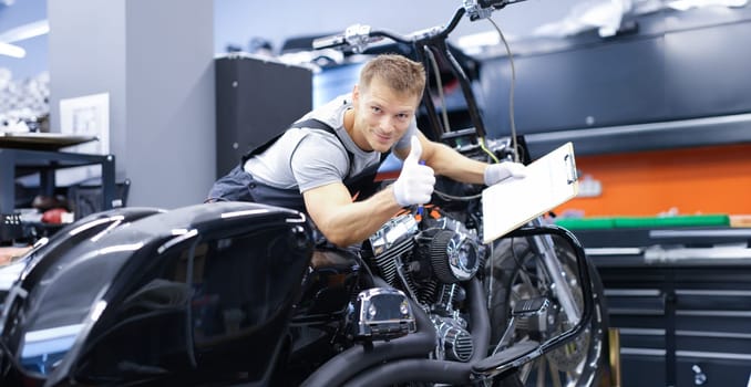 Young car mechanic is diagnosing a motorcycle and holding clipboard and thumbs up. Quality motorcycle service concept