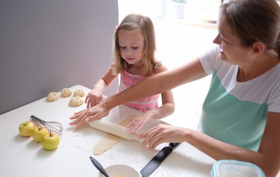 Mom and daughter rolling out flour dough at table in kitchen. Parenting children concept