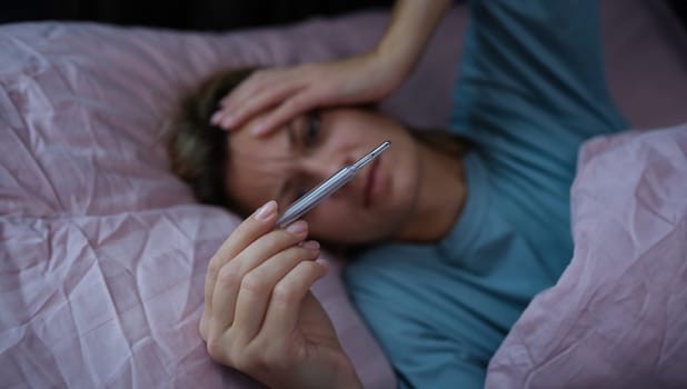Sick woman lying in bed and holding thermometer with high temperature closeup. Seasonal colds home treatment concept