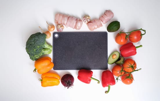 Black cutting board lying in middle of chicken meat and vegetables on table top view. Home cooking concept