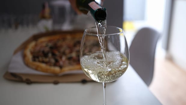 White wine pouring from bottle into glass goblet on pizza background closeup. Anniversary celebration concept