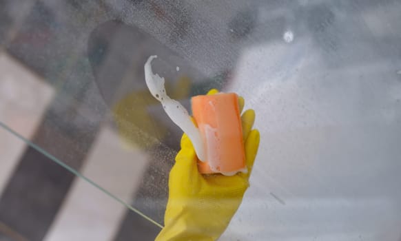 Cleaner in rubber gloves washing glass in bathroom with detergent and sponge closeup. Apartment cleaning concept