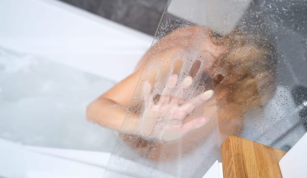 Naked woman sitting in bathtub and holding her hand on misted glass closeup. Treating stress and depression in adolescents concept