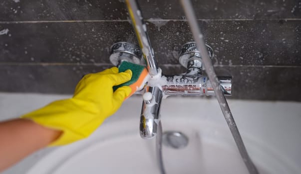 Housewife in rubber glove washing faucet in bathroom with sponge closeup. Use of household chemicals for cleaning plumbing in bathroom concept